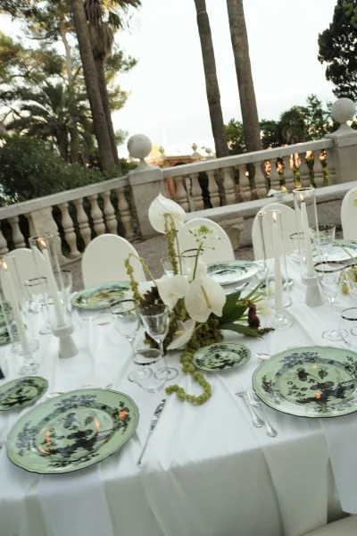 Reception tablescape with green patterned plates on white tablecloth, anthurium centerpiece and taper candles on a stone terrace with palms