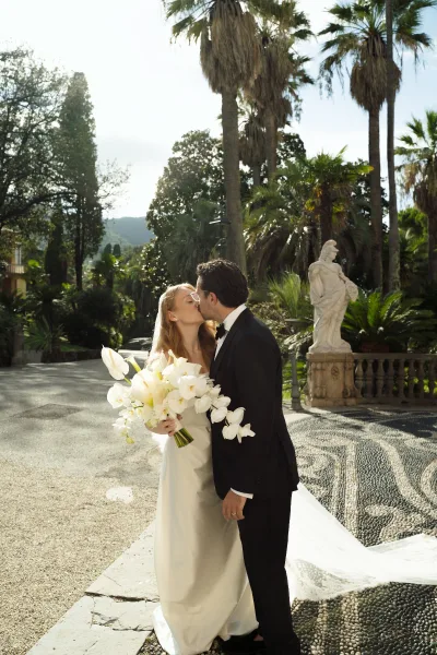 Wedding kiss portrait of bride and groom kissing, bride holding a cascading orchid bouquet on a palm-lined stone terrace with statue backdrop
