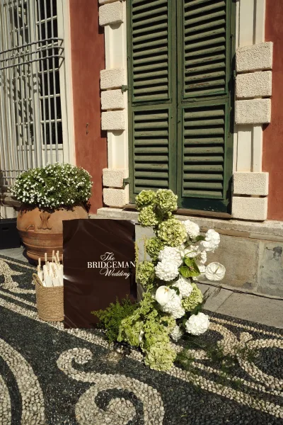 Wedding welcome sign with flowers, framed by hydrangeas and potted blooms, beside a basket of white parasols on cobblestones by a facade