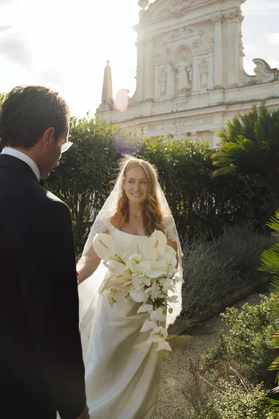 First look moment as groom in tuxedo and glasses sees bride in strapless satin dress and veil holding white orchid bouquet by stone church facade