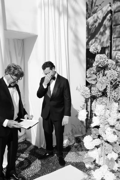 Ceremony moment as a groom in a black tuxedo wipes tears beside an officiant with microphone, framed by white draping and florals.