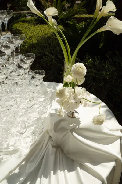 Champagne tower with champagne coupe tower glasses on a white-draped table, accented by calla lilies and lush garden greenery in sunlight