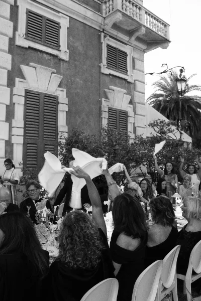 Wedding reception toast as guests cheer and wave napkins around long candlelit tables with florals in an outdoor courtyard by a historic facade