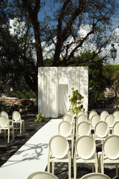 Ceremony setup for an outdoor wedding ceremony with a white aisle runner, white chairs, and a draped backdrop on a stone terrace under trees