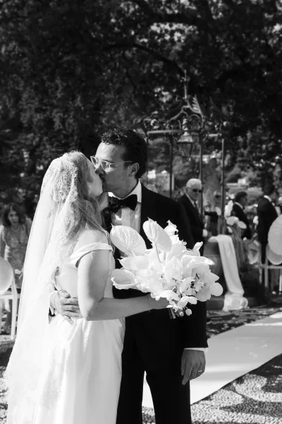 Wedding kiss as bride and groom kiss on an outdoor garden aisle, bride holding a white anthurium bouquet with lace veil flowing behind