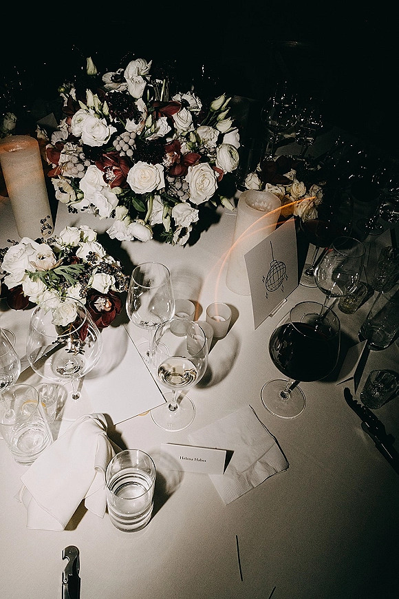 Reception tablescape with wedding table centerpiece of white roses, burgundy blooms, berry accents, pillar and votive candles in moody lighting