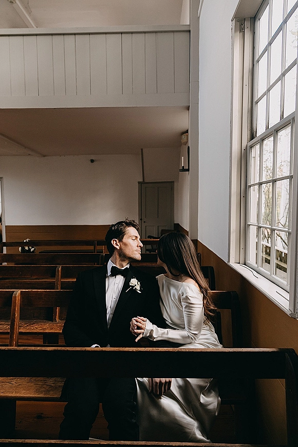 Couple portrait of bride and groom sitting in pews, holding hands as she leans on him in a church with soft window light