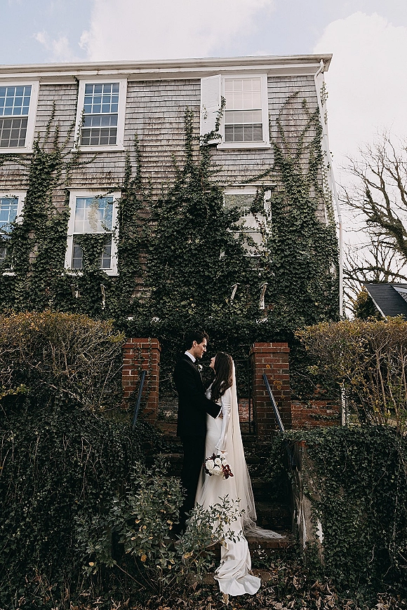 Couple portrait of bride and groom embracing on brick steps, bride holding bouquet with long veil, against ivy-covered house backdrop