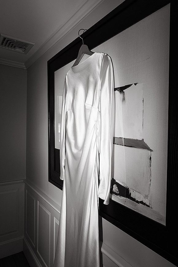 Wedding dress with long sleeves in satin hangs on a hanger in a neutral indoor hallway, framed artwork nearby and a boat neckline visible