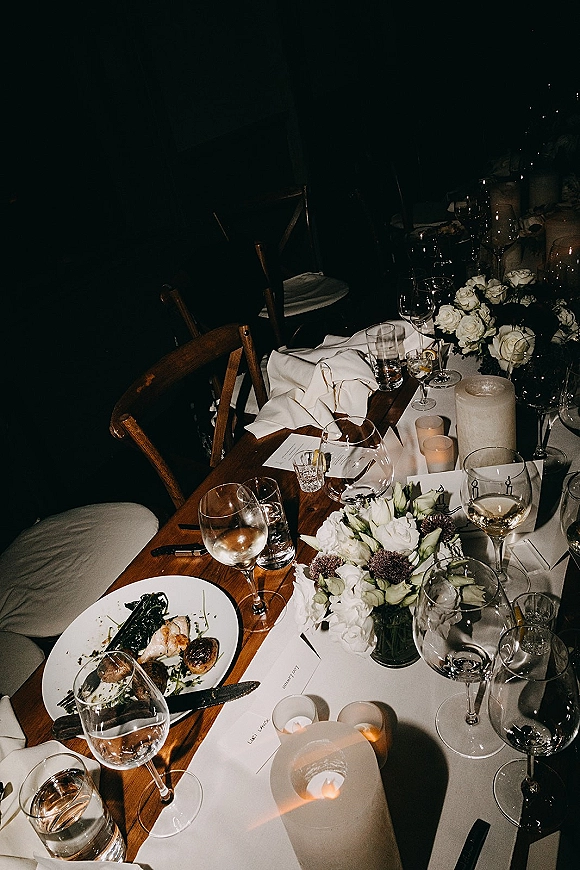 Reception tablescape with a candlelit wedding table, white rose centerpiece, pillar and votive candles, place settings on a wood runner in a dark room