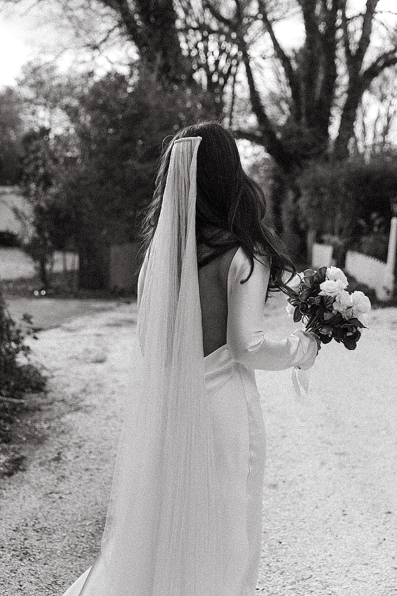 Bridal portrait of a bride from behind holding a bouquet, her long veil flowing over a strapless gown on a tree-lined garden path