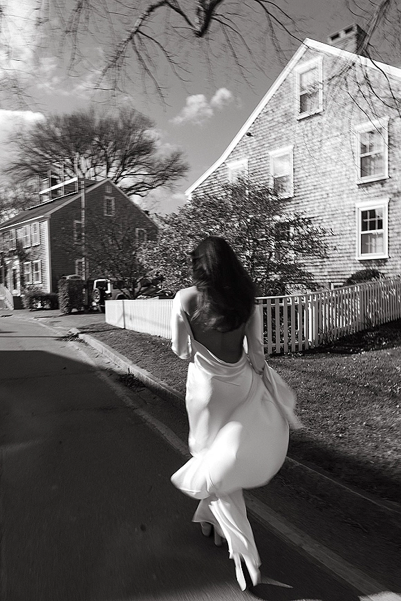 Bride portrait of a bride walking away in an open back long-sleeve gown, lifting her train on a quiet residential street with picket fence