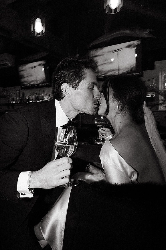 Wedding kiss as bride and groom sip champagne flutes, her veil and off-shoulder gown glowing in a bar with pendant lights and bottles
