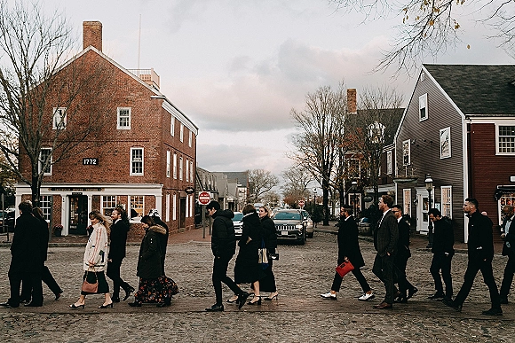Wedding guest candid of a group walking together in formal suits and long coats, one in a floral skirt, along a cobblestone street by brick storefronts