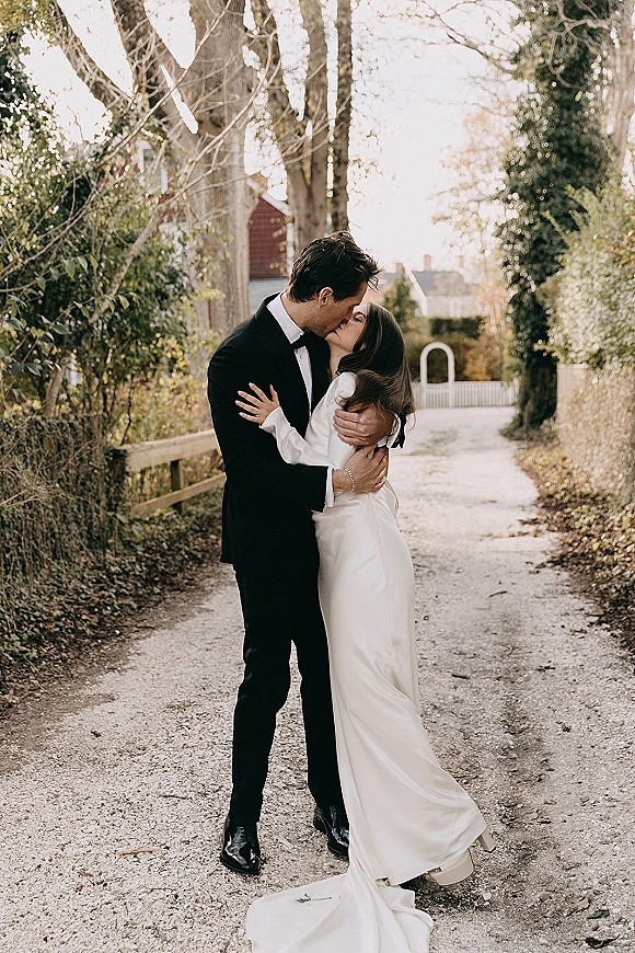 Wedding kiss as bride in long sleeve gown and groom in black tuxedo embrace on a tree-lined gravel lane by a wooden fence