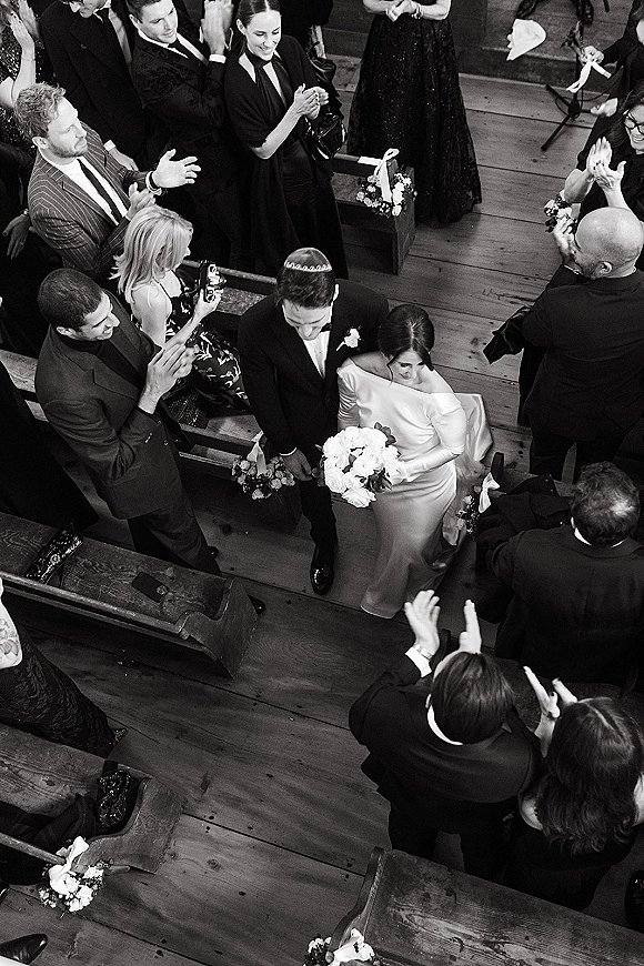 Wedding recessional as bride and groom walk the aisle, guests cheering around them, overhead black-and-white view between wooden pews with flowers