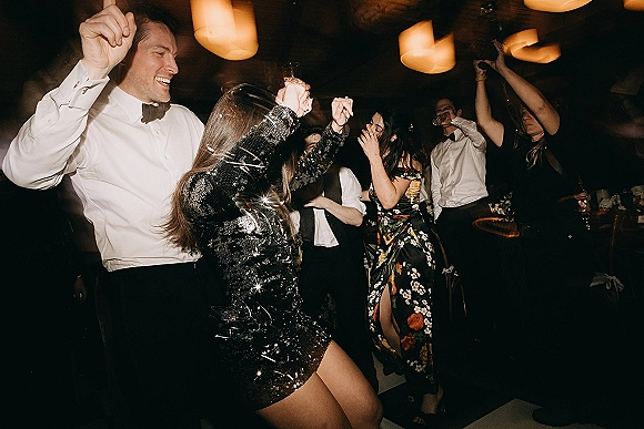 Wedding dance floor packed with guests dancing and holding drinks, with tuxedo bow tie and sequined mini dress under warm pendant lights