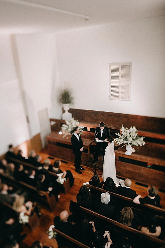 Wedding ceremony with couple exchanging vows as the officiant reads at the chapel altar, guests in wooden pews, bride’s long veil visible
