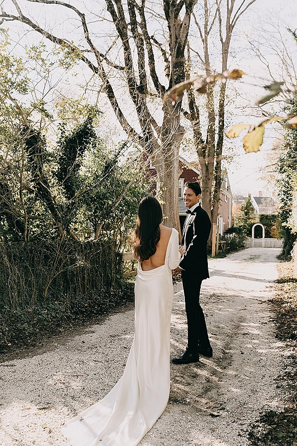 Wedding couple portrait of bride and groom holding hands on a tree-lined autumn path, her backless gown train and his black tuxedo, cottage behind