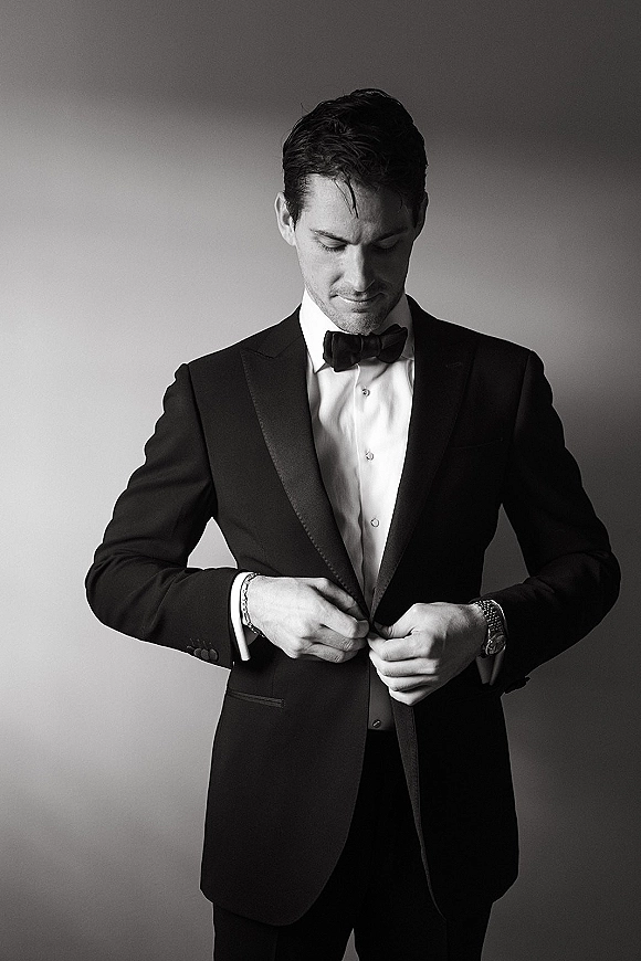 Groom portrait in a black tuxedo adjusting his jacket, showing bow tie, cufflinks, and wristwatch against a plain studio wall