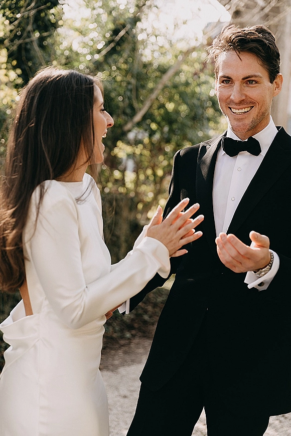 Wedding candid moment as bride laughs in an open-back gown beside smiling groom in black tuxedo, sunlit trees and greenery behind