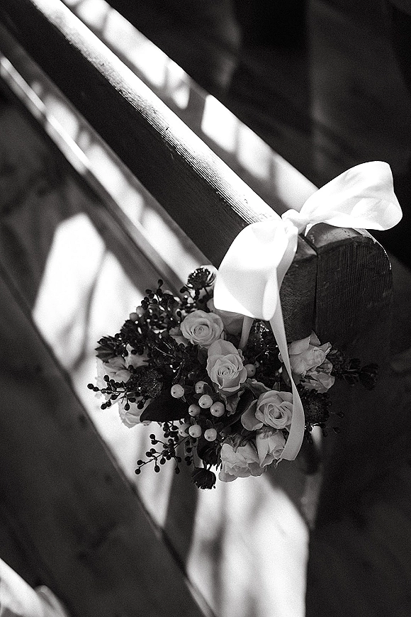 Aisle chair decor with a white ribbon bow and rose floral spray with berries and greenery on a wooden pew in striped light shadow