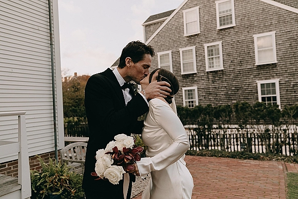Wedding kiss as groom holds bride’s face, her white rose bouquet with burgundy accents in hand, outdoors by a shingled house walkway