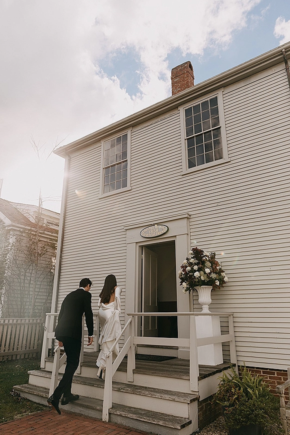 Couple portrait of bride and groom walking up wooden porch steps, her long veil trailing, toward a white clapboard venue in sun flare