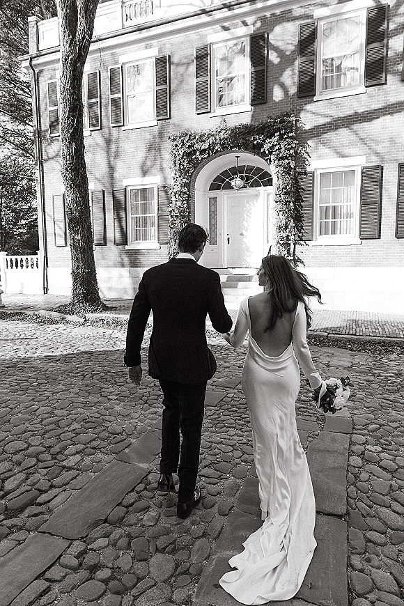 Couple portrait of newlyweds holding hands, walking away toward an ivy-covered brick doorway, bride in open-back gown with bouquet