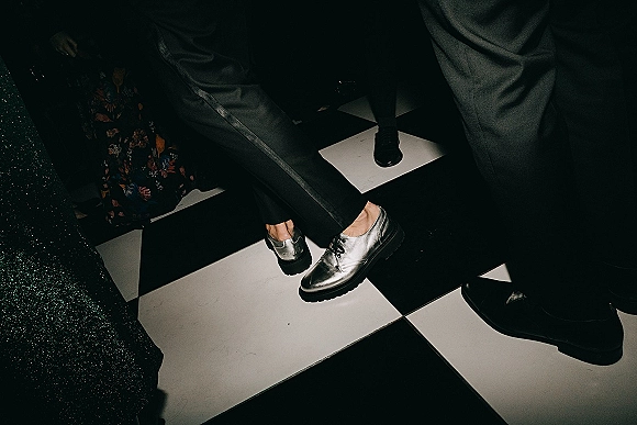 Groom shoes in metallic silver wedding shoes with black socks and suit trousers on a black-and-white checkered floor beside floral fabric