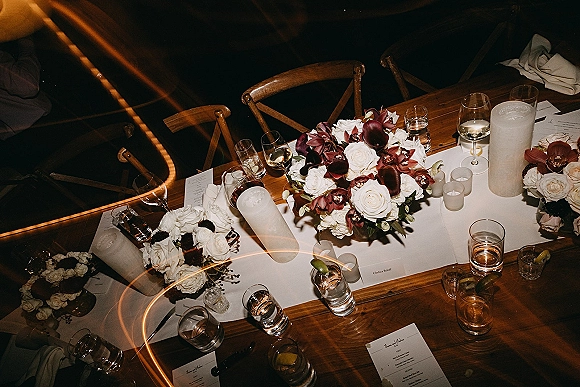 Reception tablescape with wedding head table decor on a long farm table, white runner, rose-orchid centerpiece, and pillar candles in dim lighting