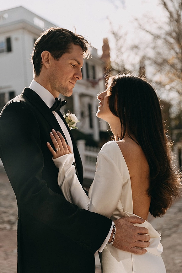 Couple portrait of bride and groom portrait embracing forehead to forehead, groom in black tuxedo with bow tie on sunny street by white building
