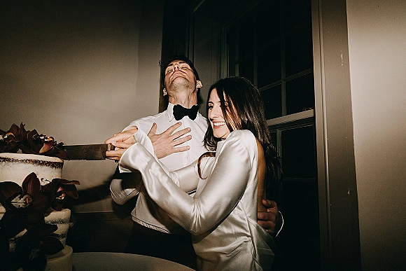 Cake cutting moment as bride and groom laugh while holding a cake knife by a floral-decorated wedding cake in a bright indoor room.