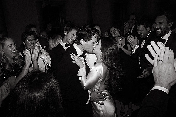 Wedding kiss moment as newlyweds embrace in a cheering crowd, groom in tuxedo holding the bride in a long sleeve satin dress at reception