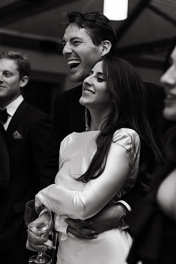 Wedding reception moment in a black and white wedding photo as the bride and groom laugh, hugging with a wine glass, guests and beams behind
