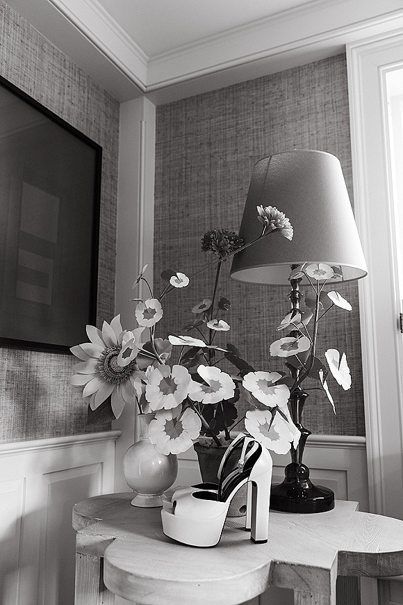 Bridal shoes, white platform wedding heels with ankle straps, styled on a side table beside a lamp and flower vase in window light