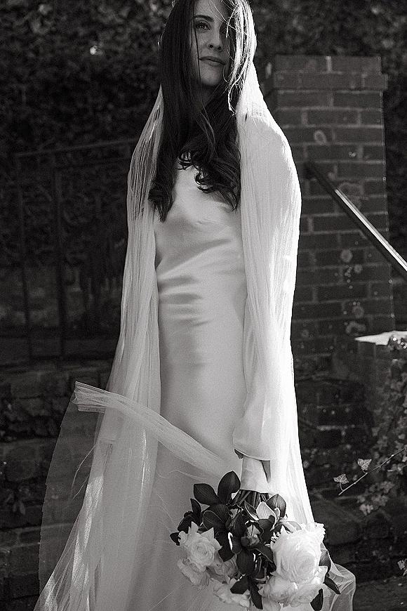 Bridal portrait in black and white of a bride in a wedding dress with long veil, holding a greenery bouquet on outdoor brick steps by foliage