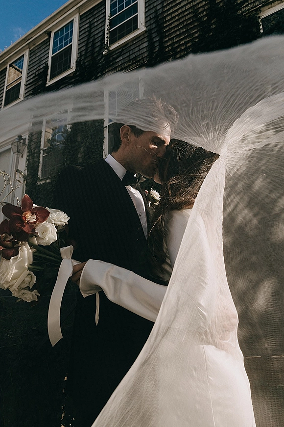 Wedding kiss portrait of bride and groom kissing as veil blows in the wind, bride holding white rose and orchid bouquet outside ivy-covered house