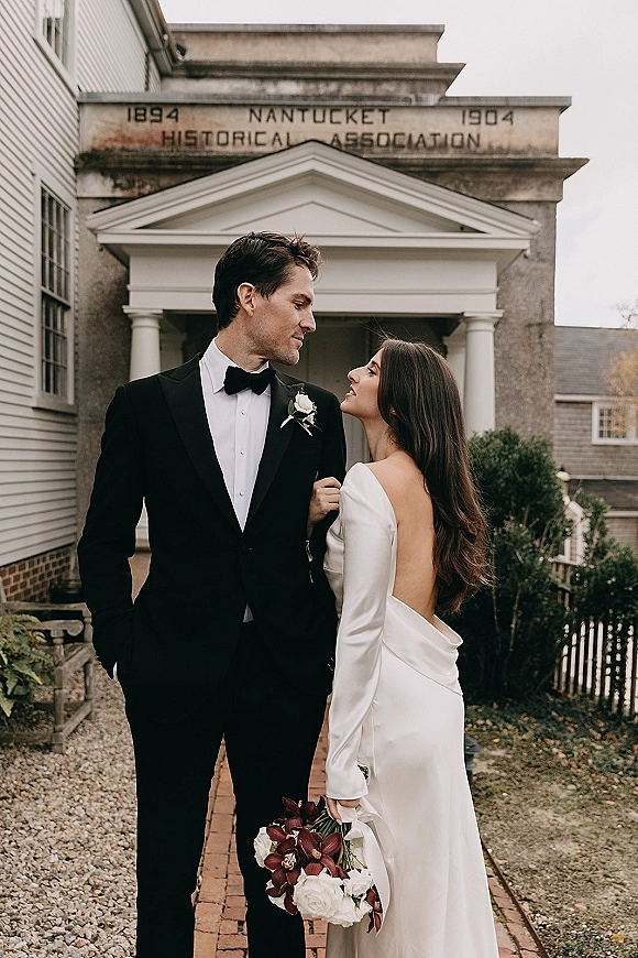 Couple portrait of bride looking up at groom in black tuxedo, holding orchid bouquet on brick walkway by historic columns under overcast sky