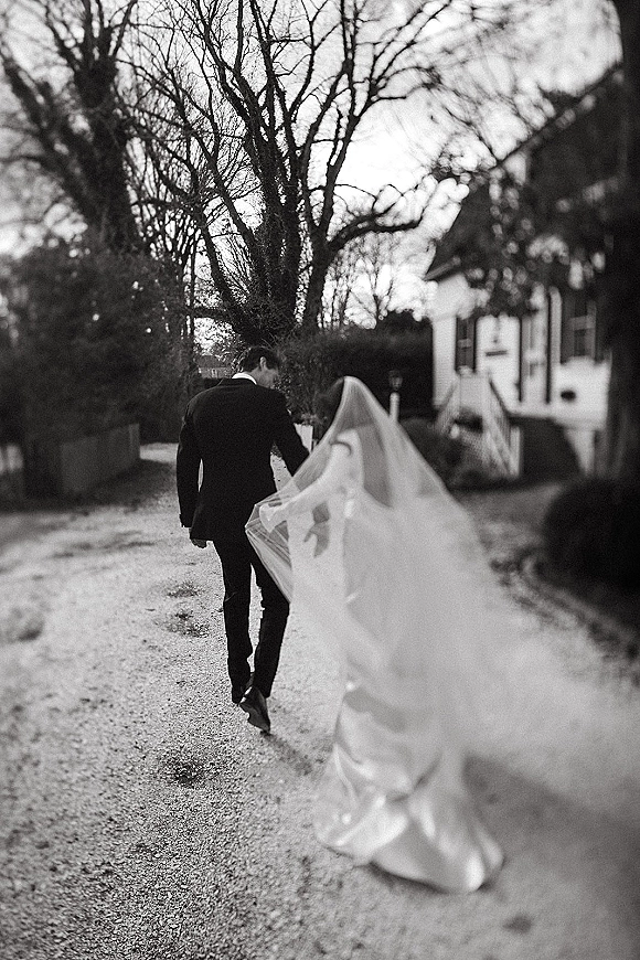 Newlywed walkaway as bride and groom walking away hand in hand, her veil blowing over a long dress train on a gravel driveway by a house