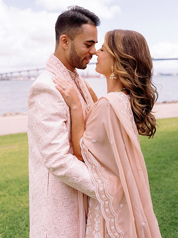 Couple portrait of a South Asian couple portrait touching foreheads, bride’s hand on groom’s embroidered sherwani by a waterfront railing under clouds