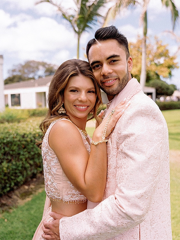 Couple portrait of bride and groom embrace, showcasing embroidered bridal outfit and pink sherwani on a palm-lined lawn under clouds