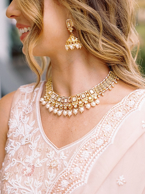 Bridal jewelry with a gold pearl necklace and chandelier earrings resting over an embroidered blouse, softly lit against blurred greenery outdoors