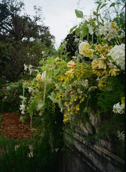 Wedding floral installation with lush greenery garland and lilies cascading over a stone wall in a garden setting under cloudy sky