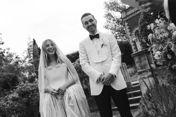 Couple portrait in a black and white wedding photo, bride laughing in long veil beside groom in white tuxedo on stone steps by brick building