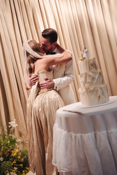 Wedding kiss as the bride in a veil and strapless gown leans into the groom in a white tux beside a three-tier cake with topper and draped curtains behind
