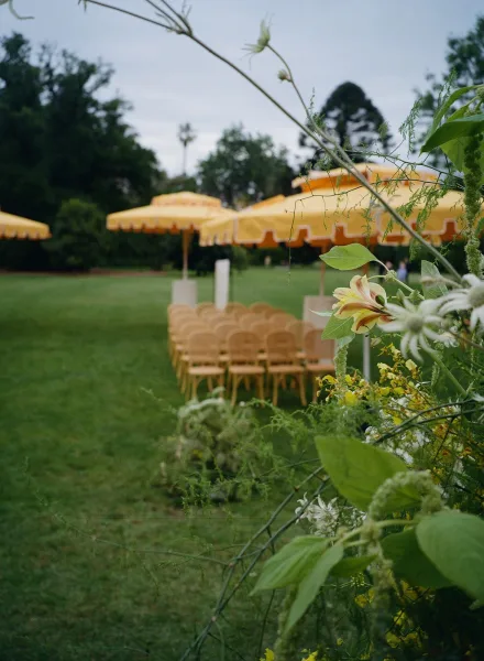Outdoor ceremony setup with yellow striped umbrellas shading rows of chairs, framed by wildflower lilies and greenery on a lawn under cloudy skies