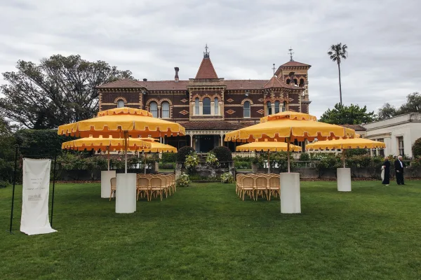 Outdoor ceremony setup with wedding ceremony umbrellas and chair rows, yellow patio umbrellas framing a lawn aisle before a brick building
