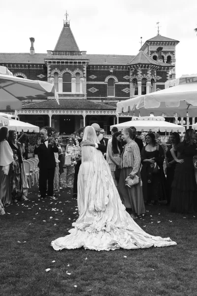 Wedding ceremony moment outdoors as bride walks down the aisle with a long veil and bouquet toward groom by a historic veranda facade