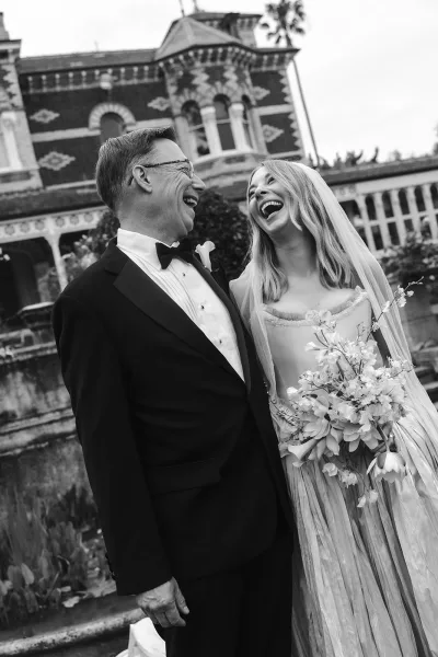 Couple portrait in a black and white wedding portrait style, bride and groom laughing as she holds a bouquet by a historic stone wall